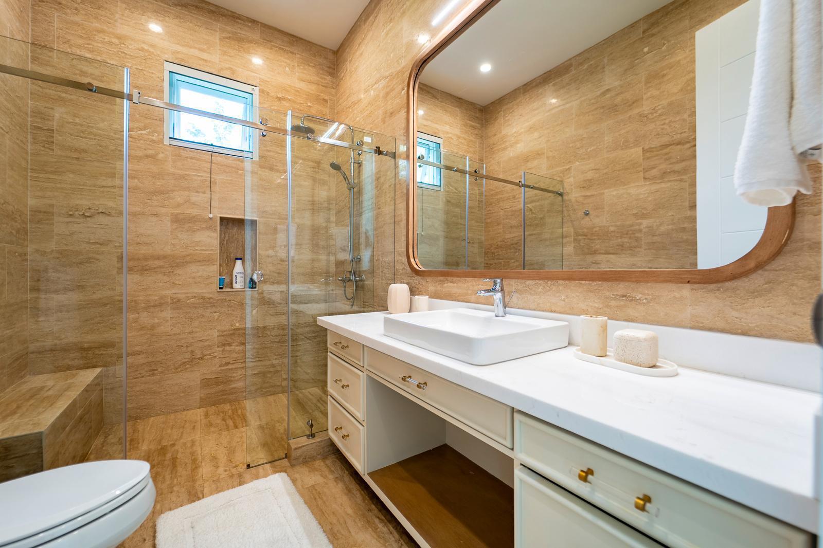 Tan stone-tiled bathroom with a glass shower, built-in bench, and long white vanity.