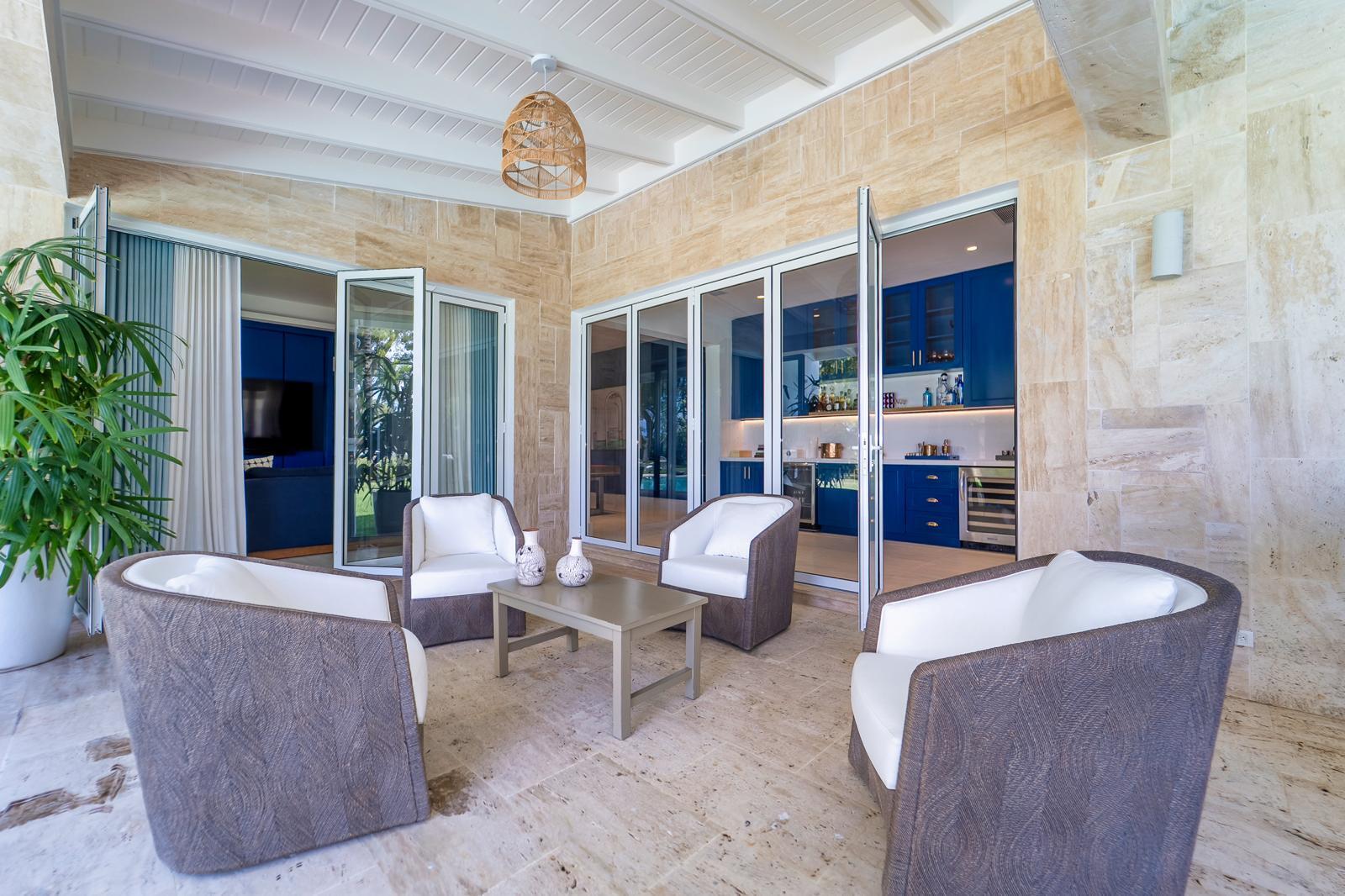Lounge with white-cushioned wicker chairs and a blue-cabinet kitchenette beyond glass doors.