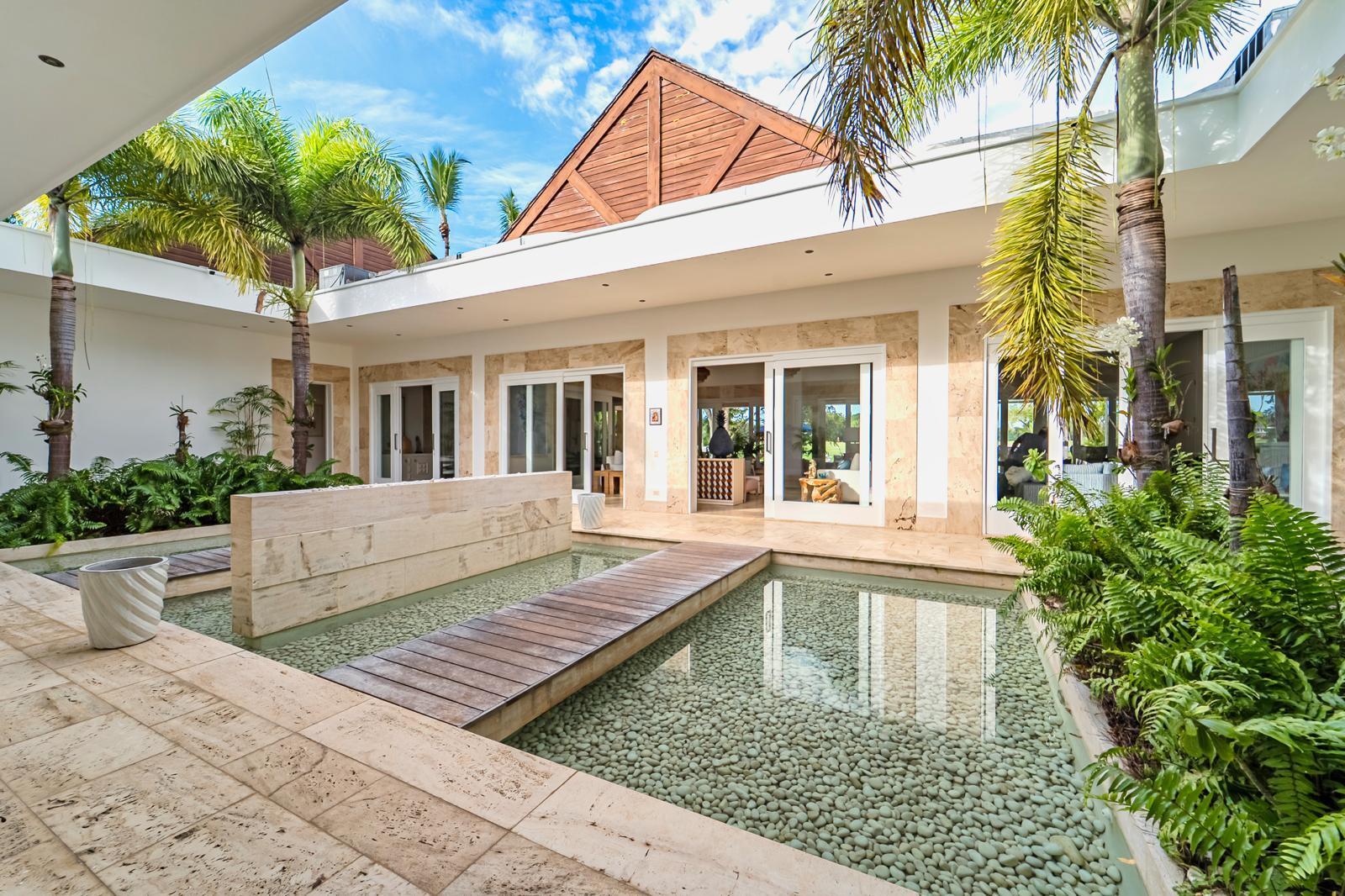 Modern home courtyard with a pebble-bottom reflecting pool, wooden walkway, palms, and stone-clad walls.