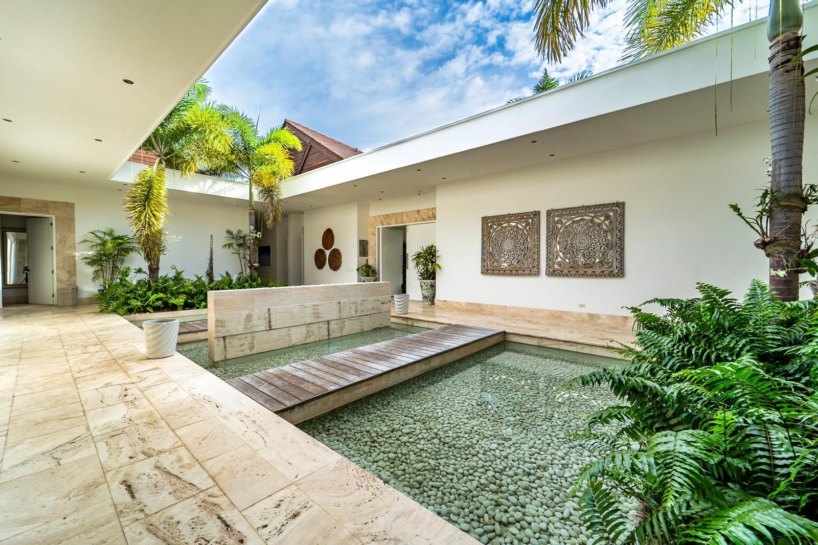 Open-air courtyard with a pebble-bottom water feature, wooden walkway, and tropical plants.