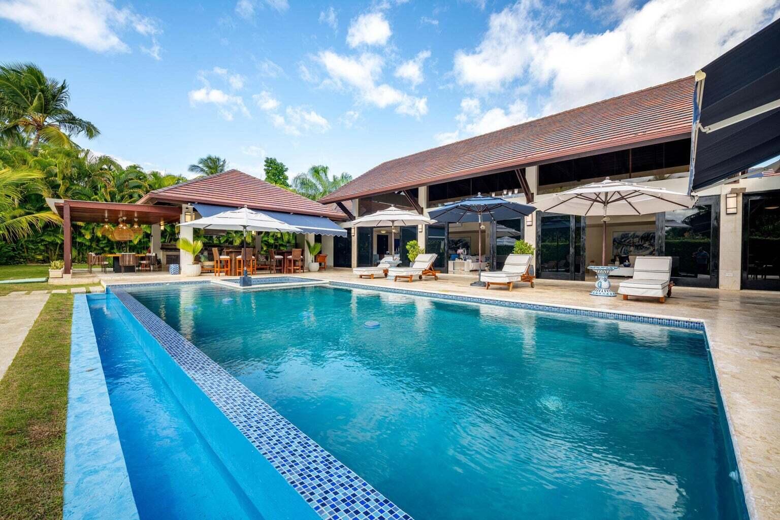 Tropical home pool with a covered dining area and umbrella-shaded lounge chairs.