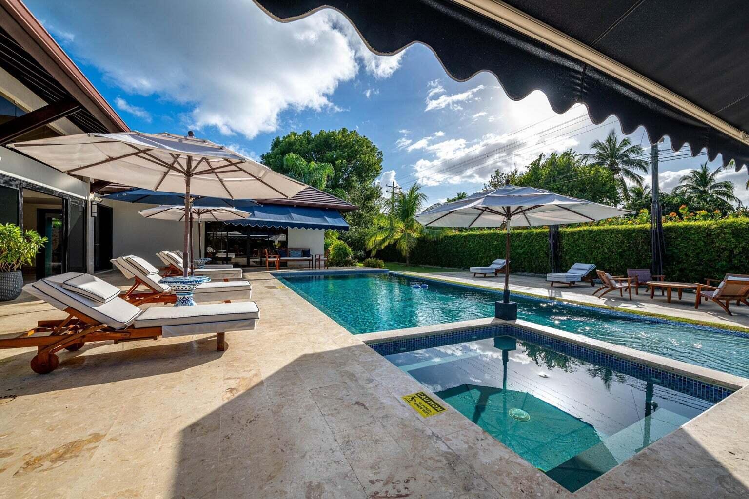 Outdoor pool and stone patio with shaded loungers, umbrellas, palms, and a clipped hedge.