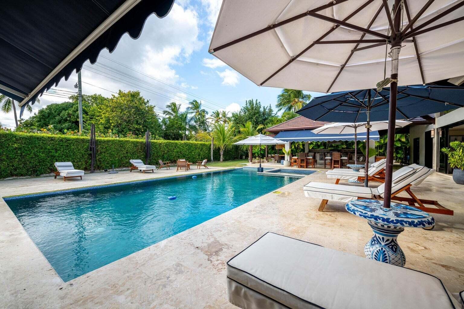 Swimming pool beside a stone patio with loungers, large umbrellas, and a covered dining area.