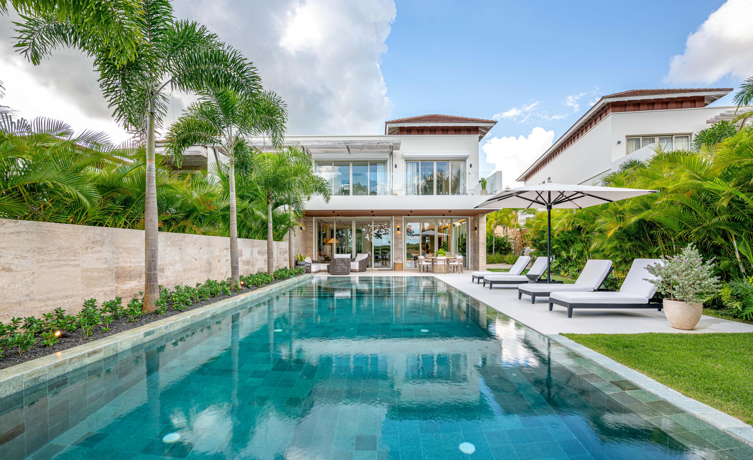 Modern white residence behind a long turquoise pool, with palm trees and poolside loungers.