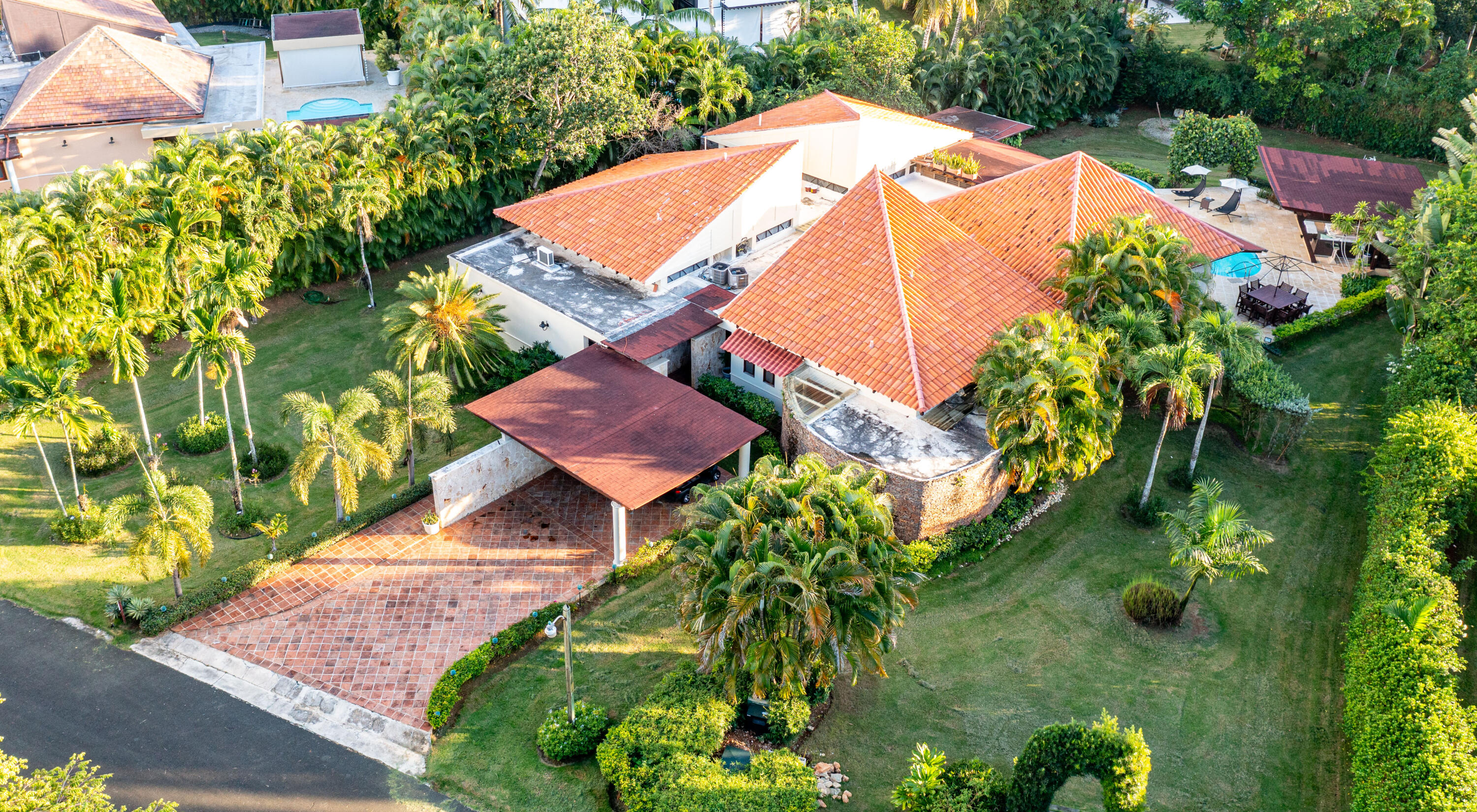 Aerial view of a terracotta-roofed house among palms, landscaped lawns, a tiled patio, and a small pool.