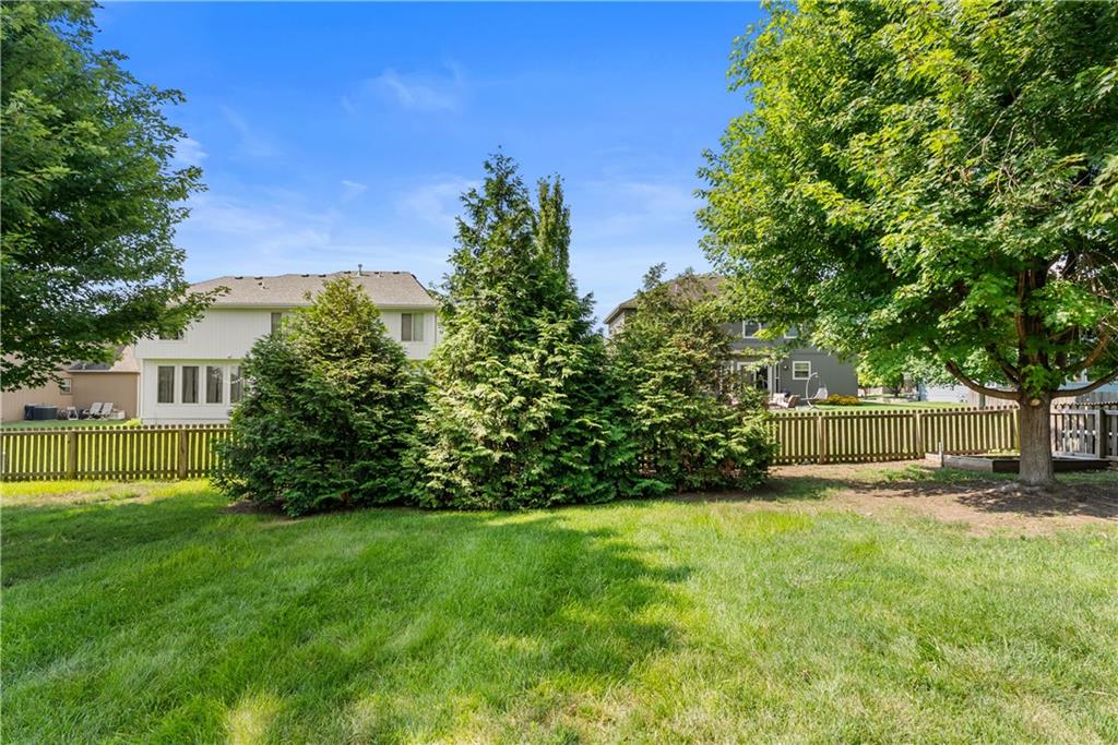 Backyard lawn bordered by mature trees, shrubs, and a wooden picket fence, with neighboring homes beyond.
