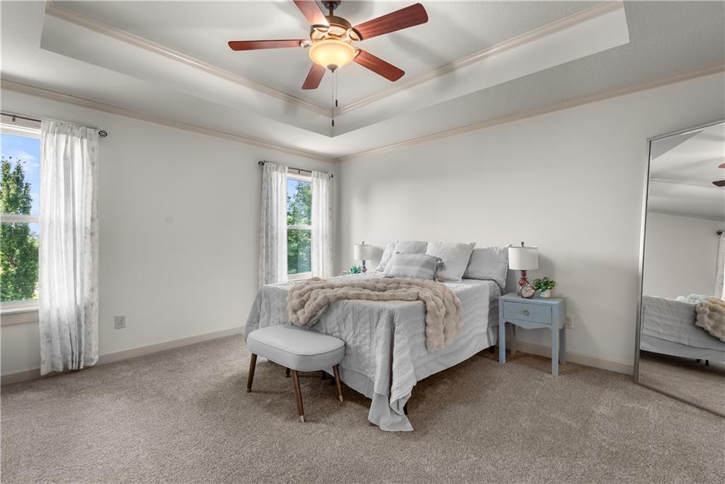 Furnished bedroom with a tray ceiling, ceiling fan, two curtained windows, and a full-length mirror.