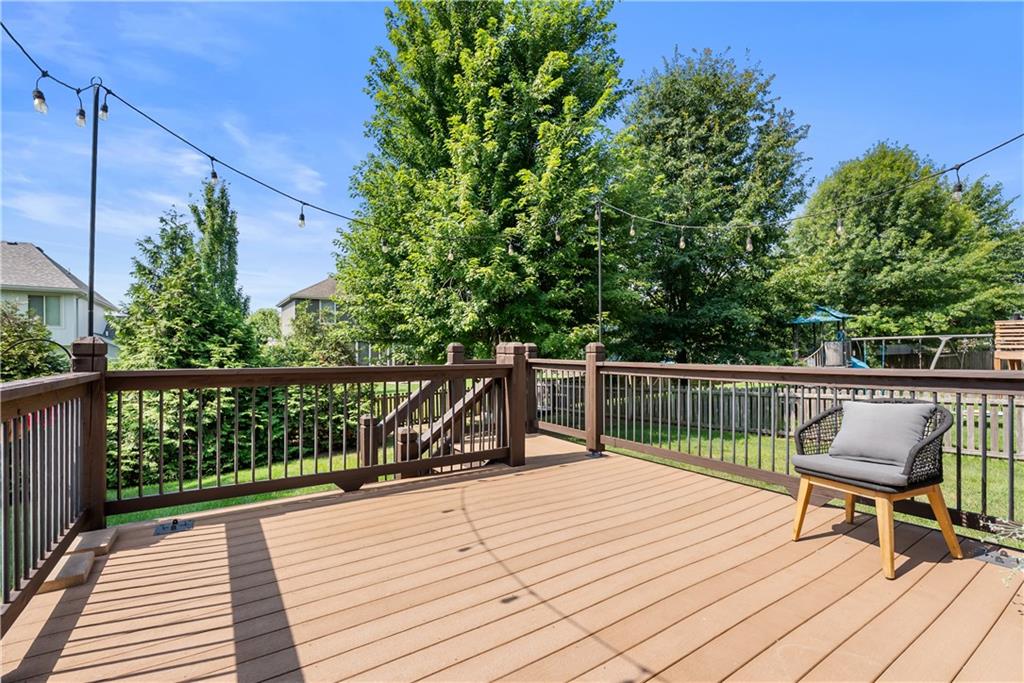 Backyard deck with dark railings, overhead string lights, and a cushioned chair among leafy trees.