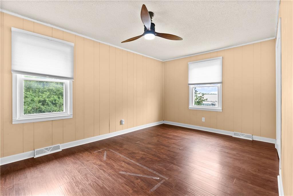 Empty room with wood flooring, tan paneled walls, two windows, and a ceiling fan.