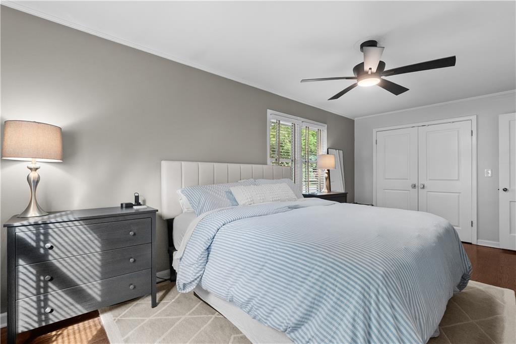 Bedroom with pale striped bedding, a white upholstered headboard, and sunlight through window blinds.
