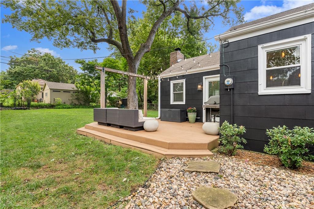 Dark-sided house with a raised wooden deck and seating beside a leafy backyard tree.