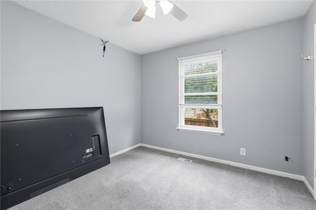 Empty gray bedroom with carpet, a ceiling fan, and a window overlooking a fenced yard.