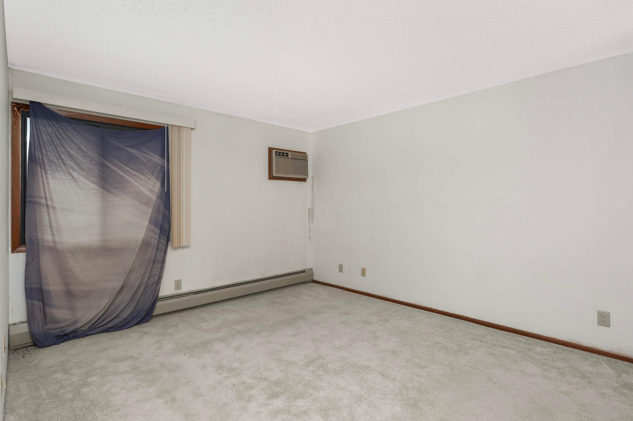 Empty carpeted room with a curtained window, wall air conditioner, and baseboard heater.