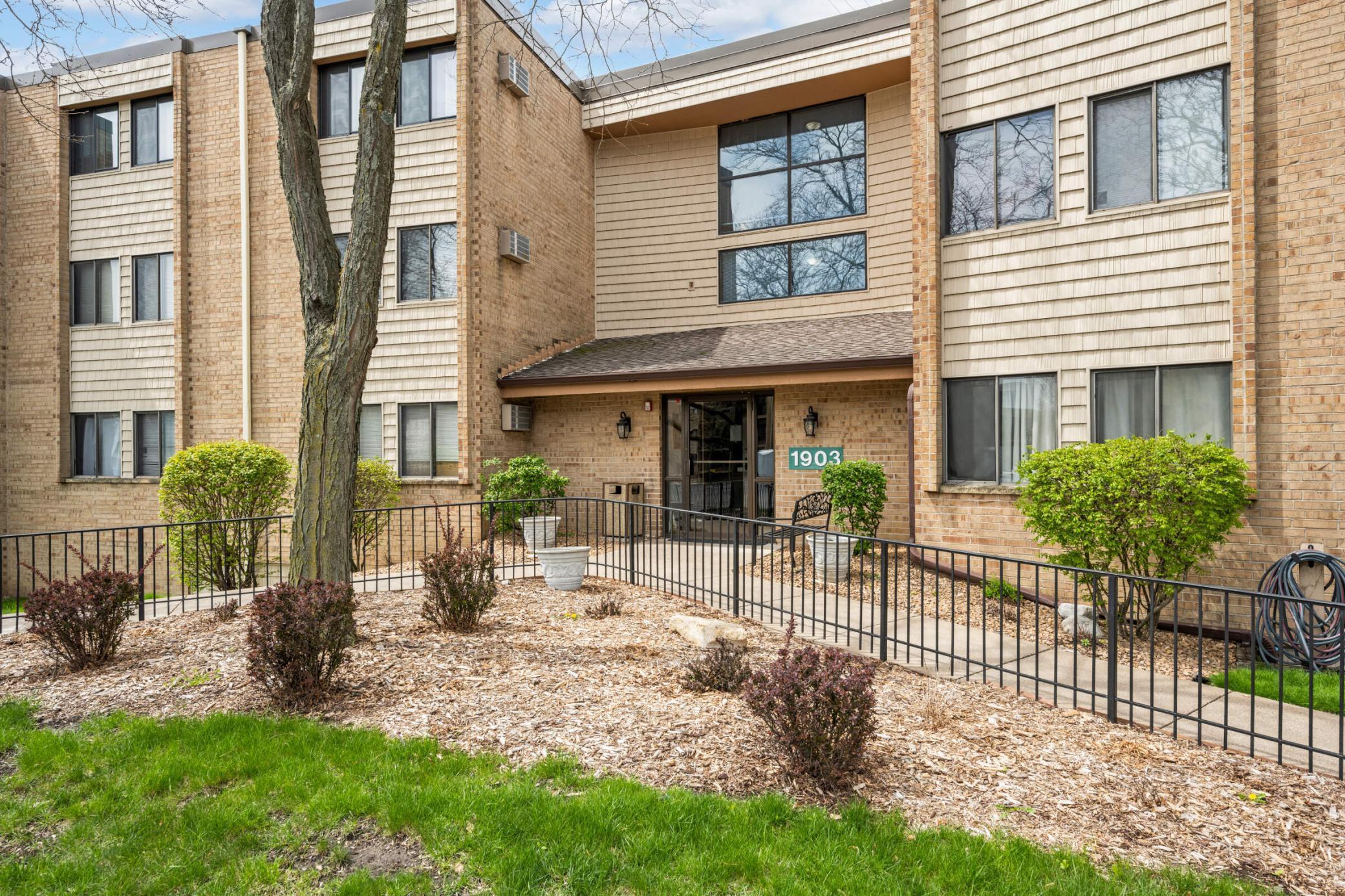 Condominium building entrance marked 1903, with brick walls, landscaped beds, and a front walkway.