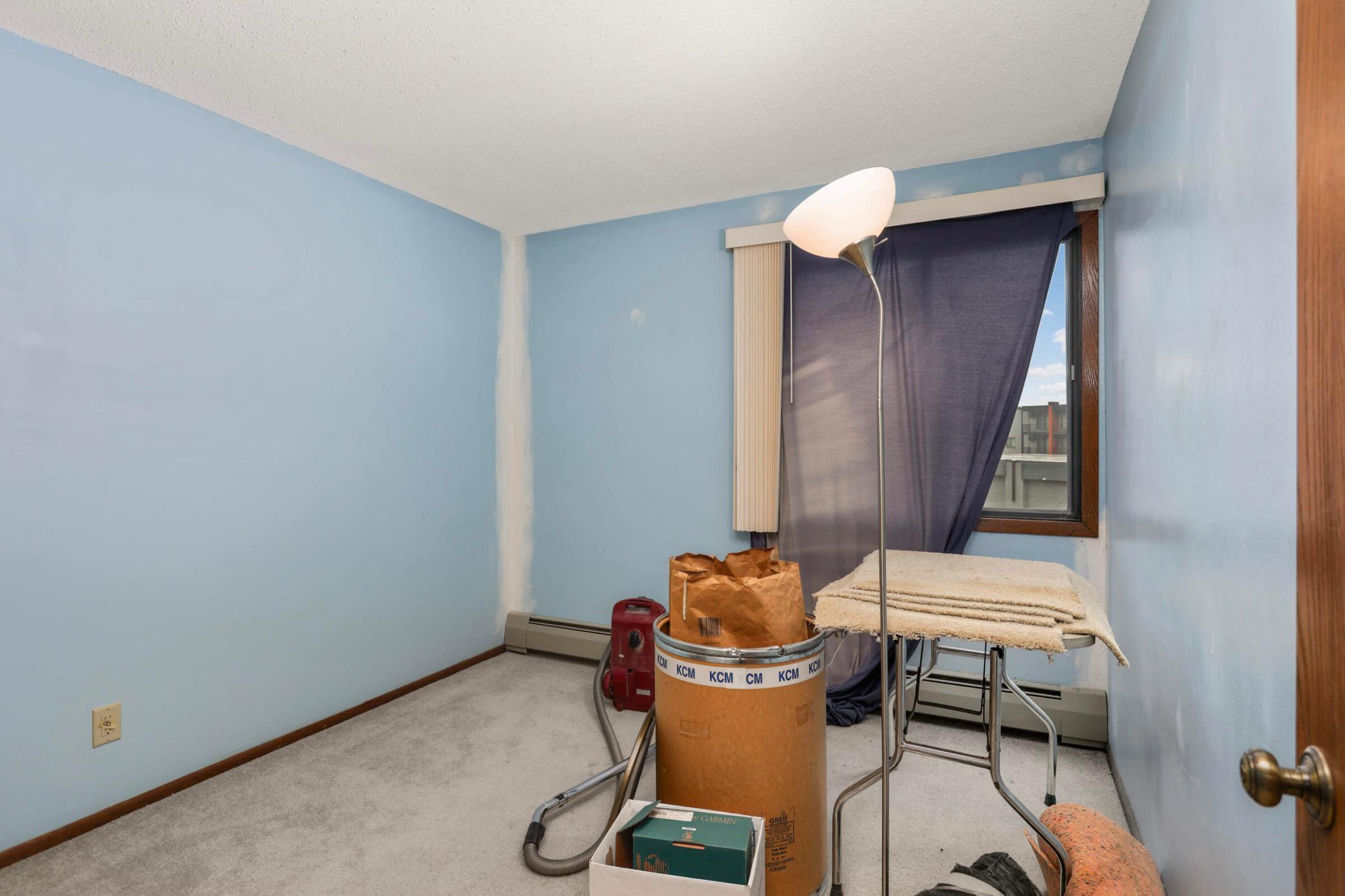 Small blue-walled room with carpet, a curtained window, floor lamp, and stored household items.