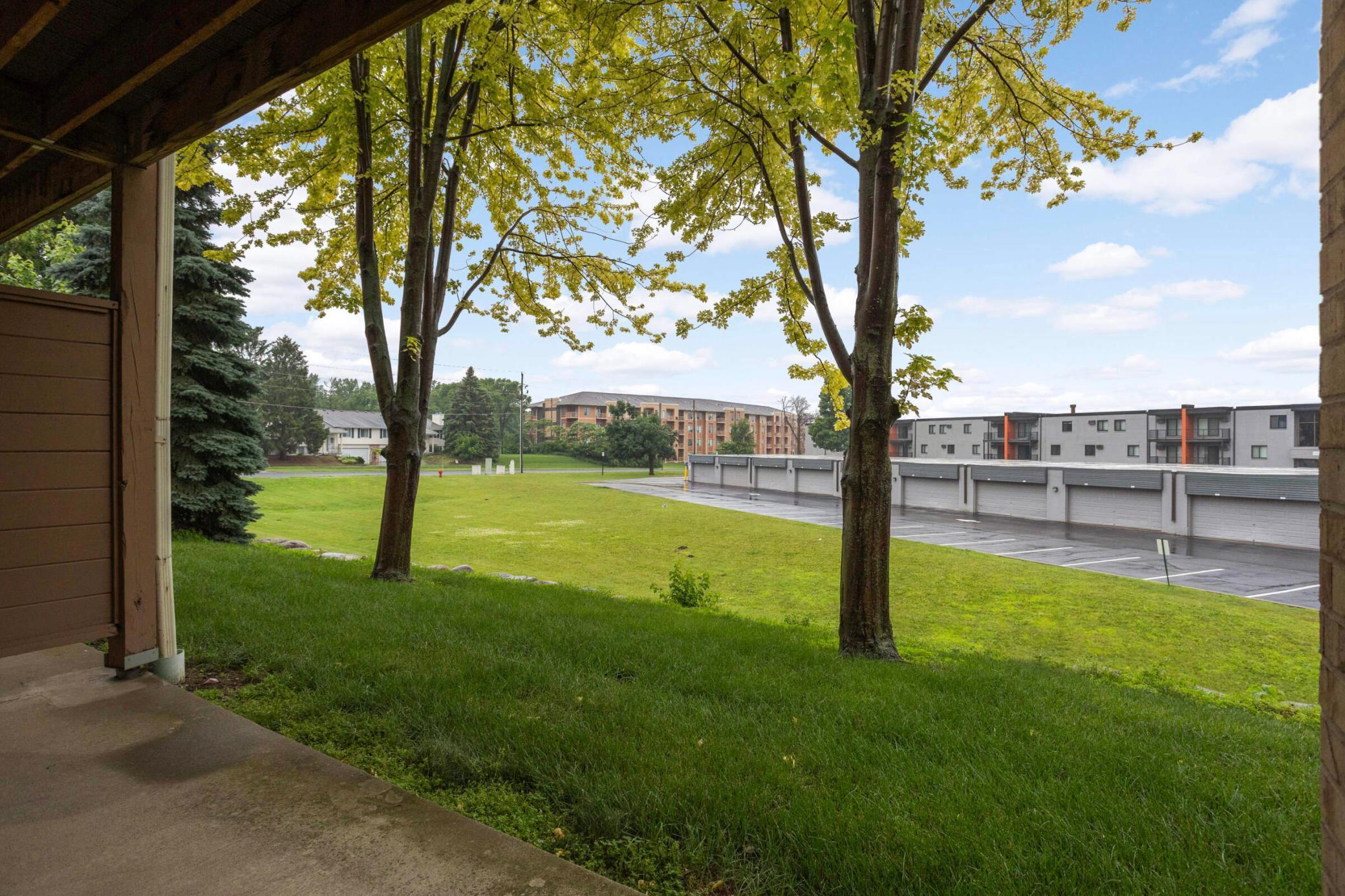 Patio view across a lawn and mature trees toward apartment buildings and parking garages.