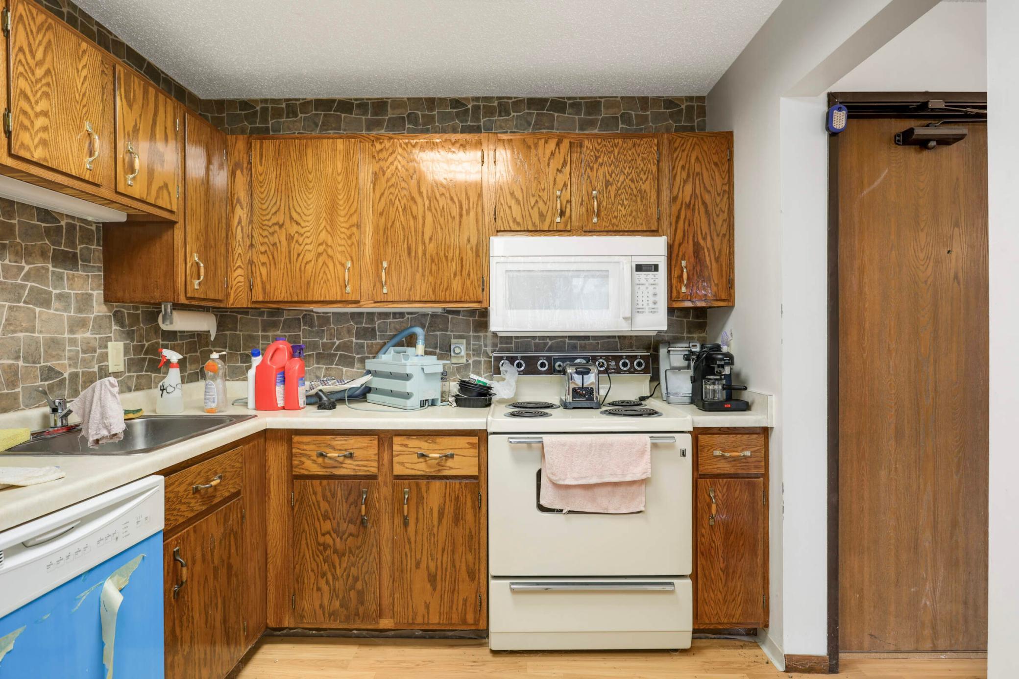 Kitchen with oak cabinets, stone-pattern backsplash, white appliances, and crowded counters.