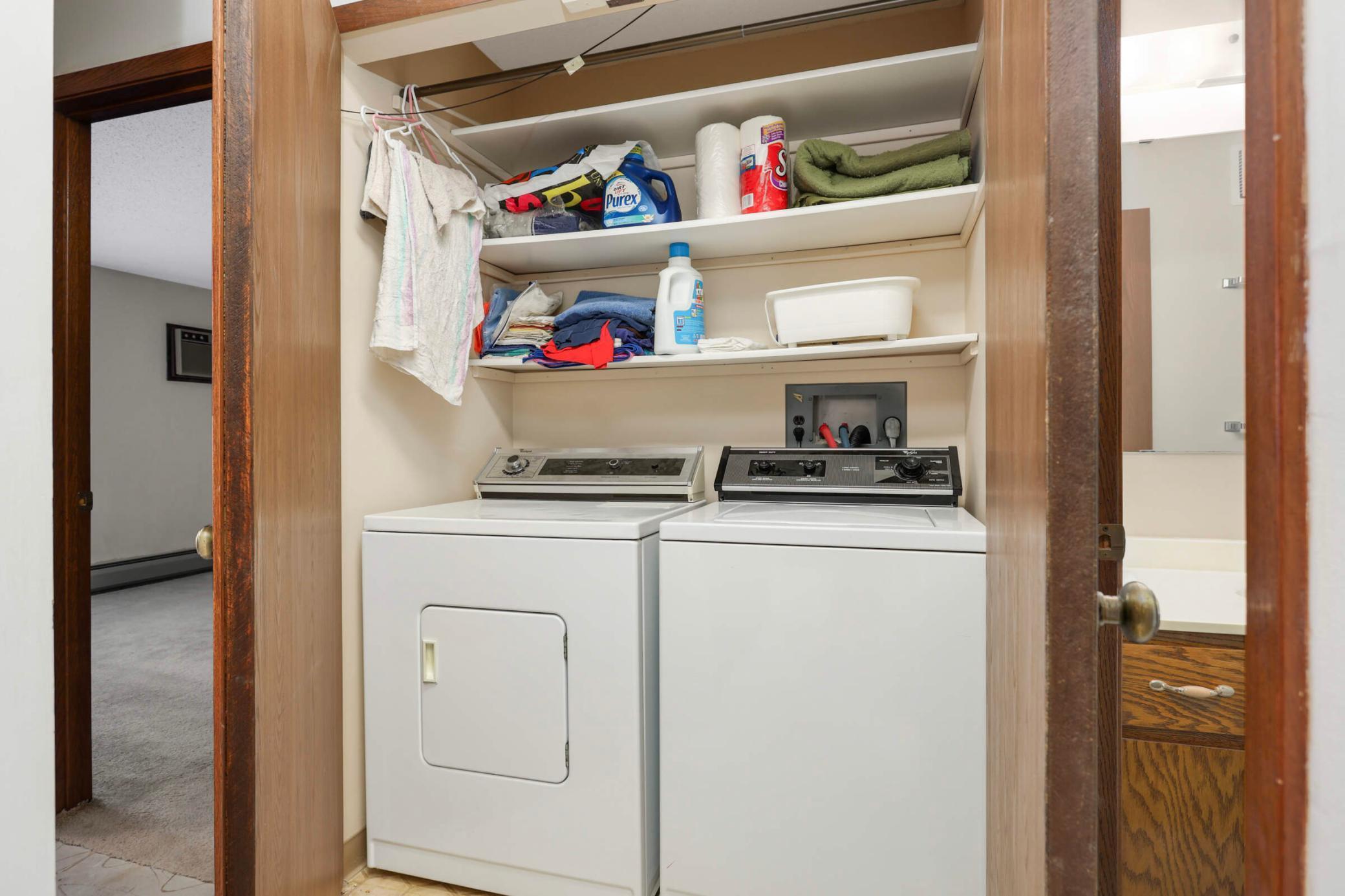 Laundry closet with a white washer and dryer beneath shelves stocked with supplies and towels.