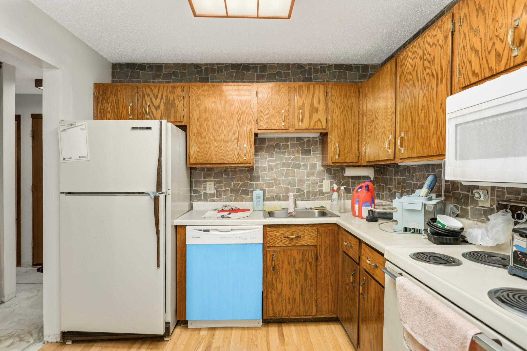 Kitchen with oak cabinets, stone-pattern backsplash, white appliances, and cluttered counters.