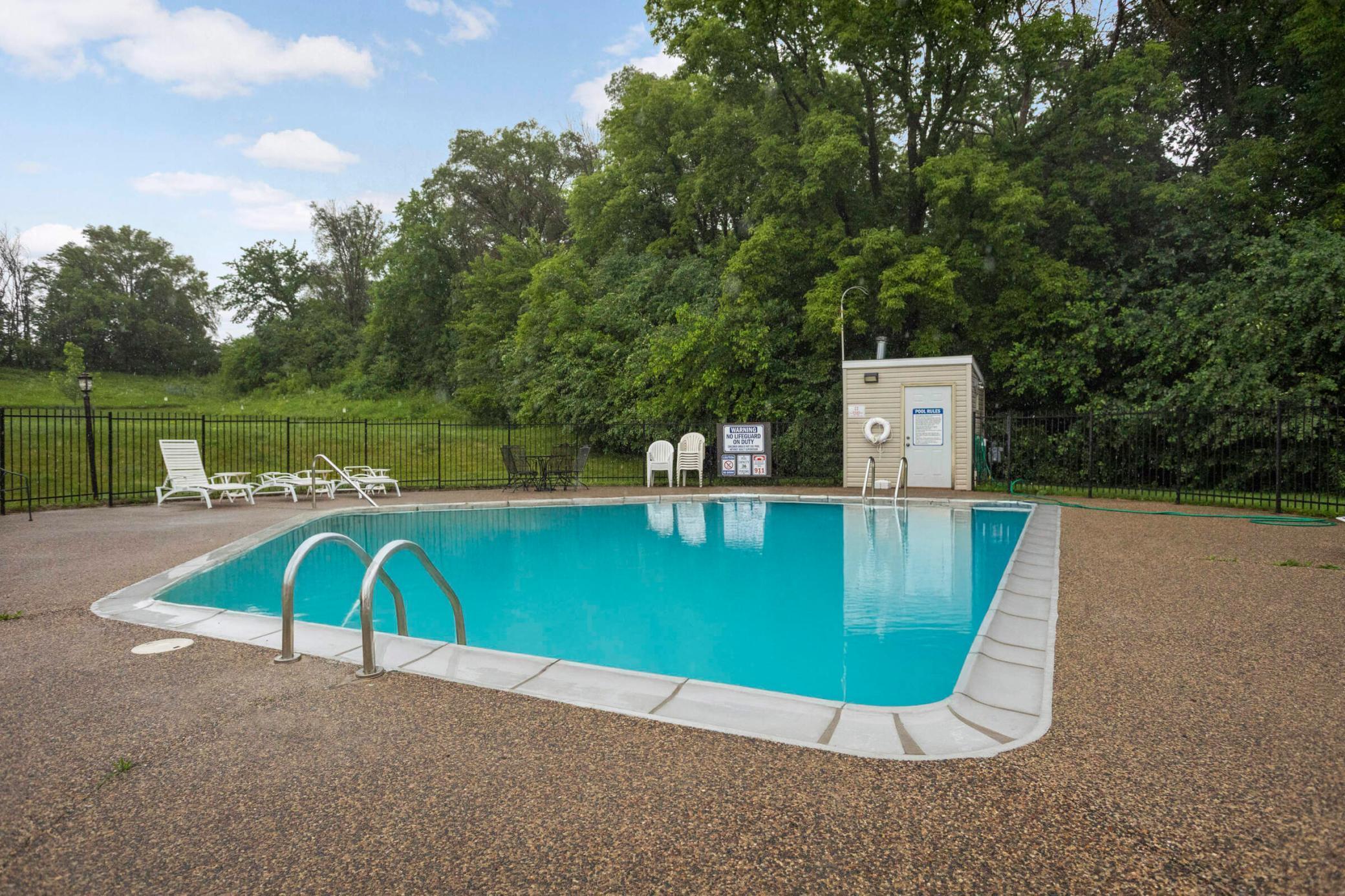 Outdoor swimming pool with a metal ladder, lounge chairs, and a tree-lined fence.