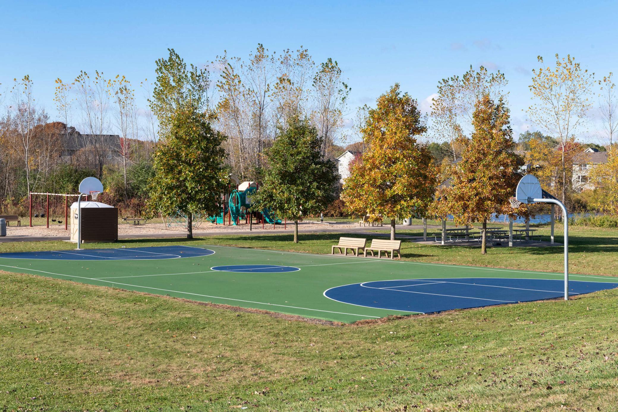 Outdoor basketball court beside a playground and picnic tables, surrounded by autumn trees.