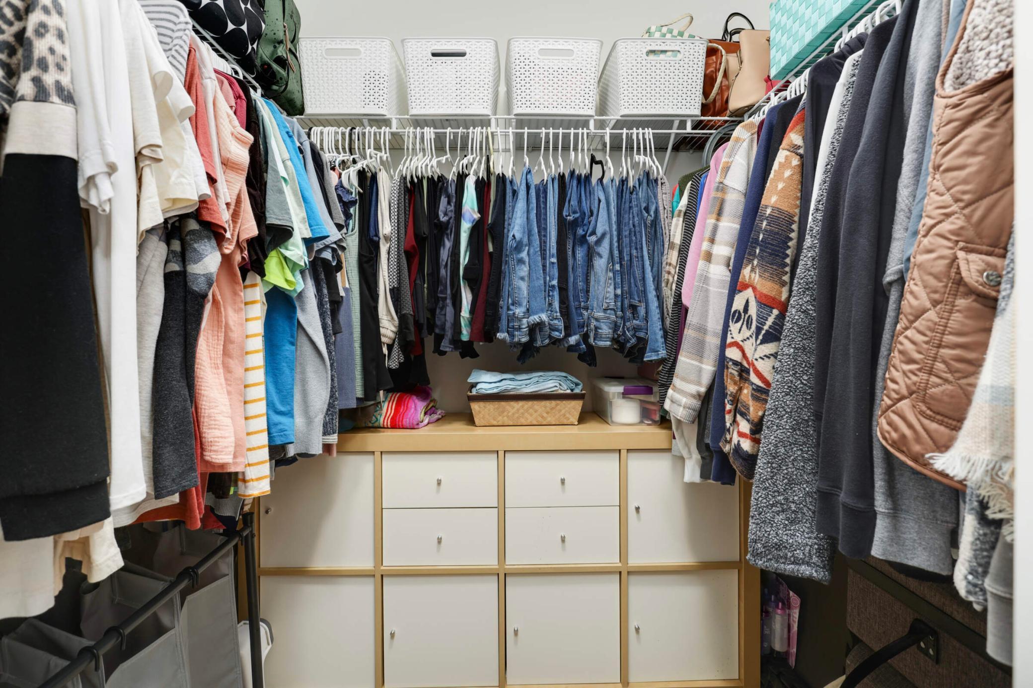 Walk-in closet with hanging clothes, upper storage baskets, and built-in white drawers and cabinets.