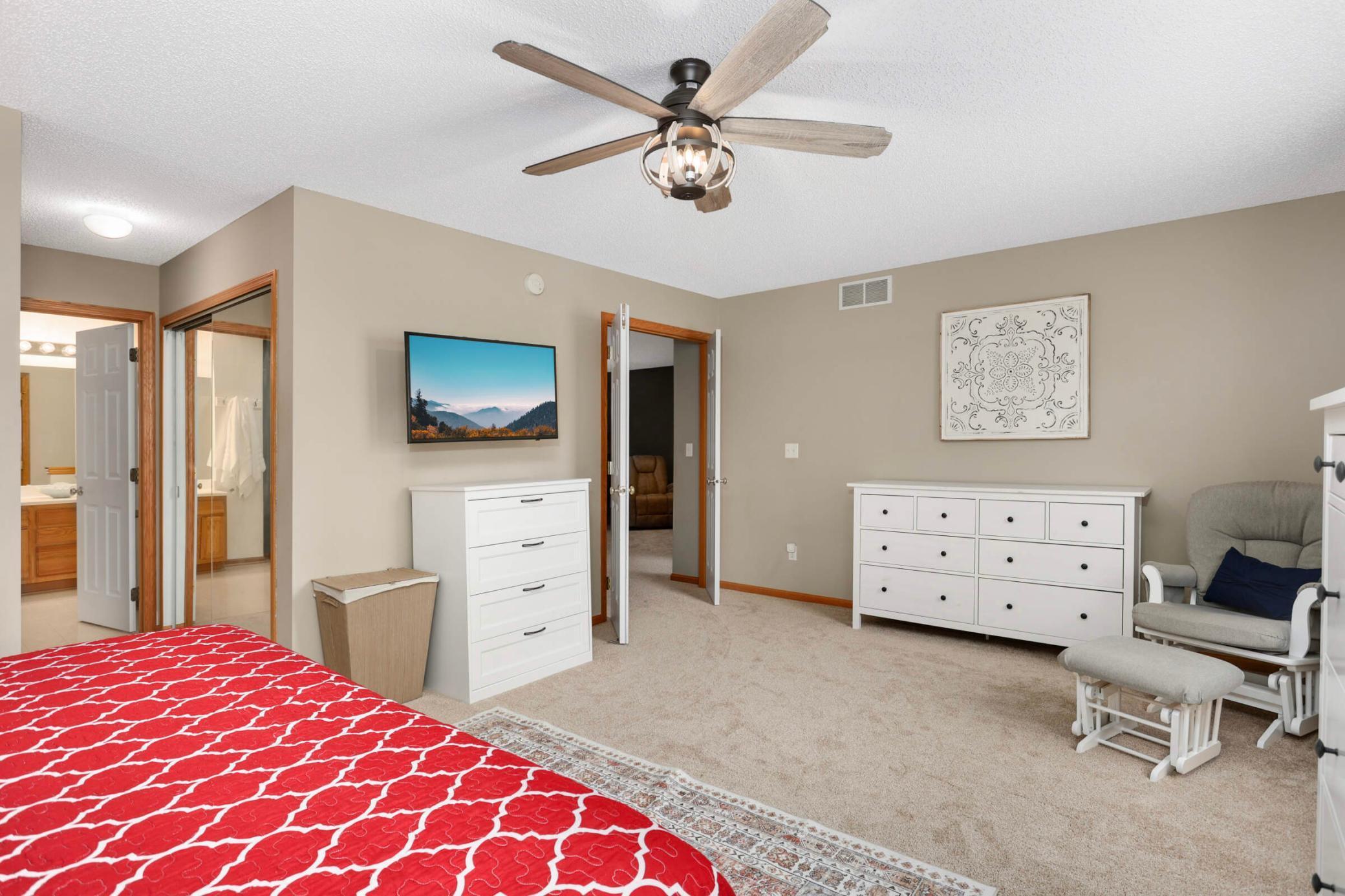 Furnished bedroom with a red patterned bedspread, white dressers, and a ceiling fan.