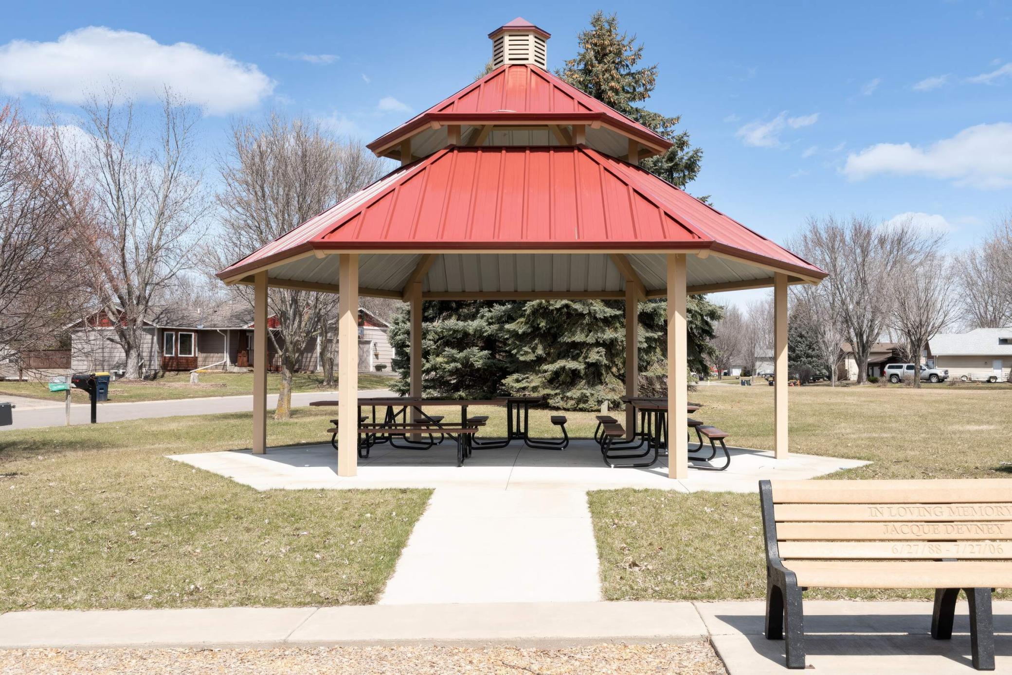 Community gazebo with a red metal roof, picnic tables, and a memorial bench nearby.