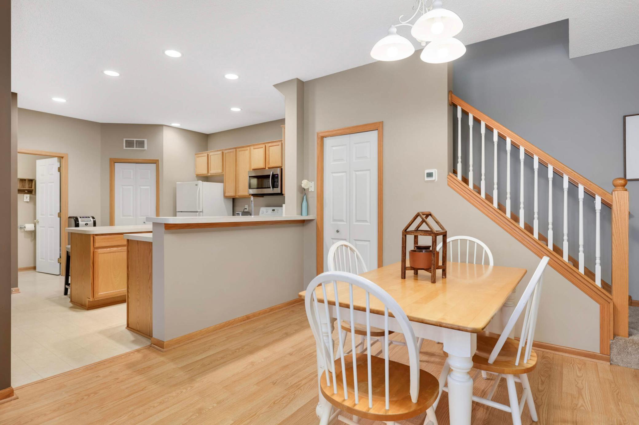 Dining area with a light wood table, white chairs, open kitchen, and wood-railed staircase.