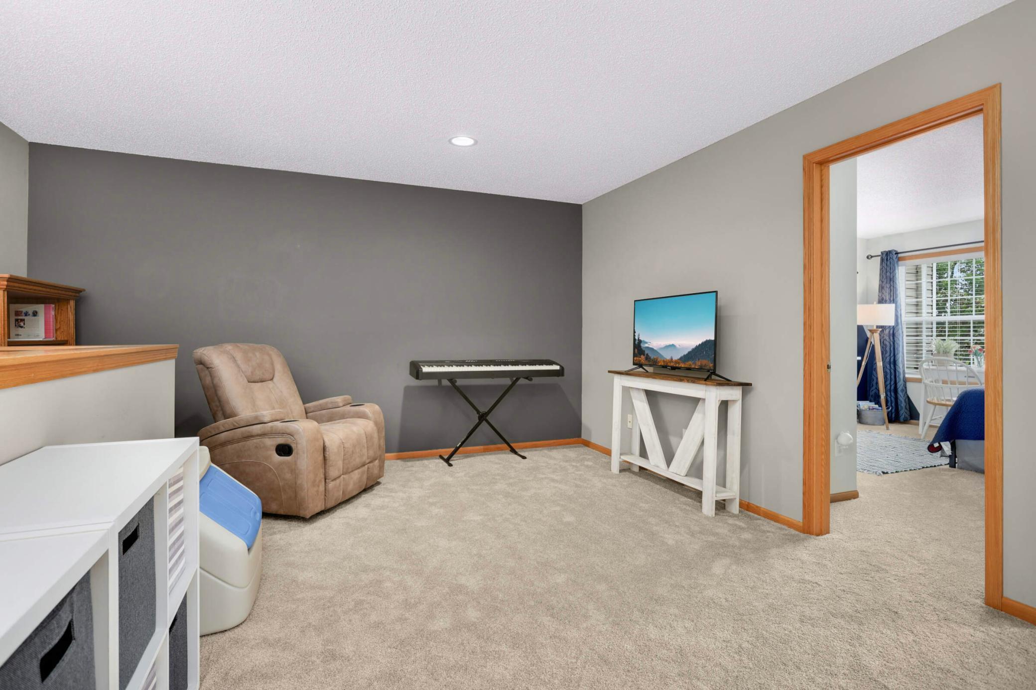 Carpeted living room with gray accent wall, recliner, keyboard, and TV beside a wood-trimmed doorway.