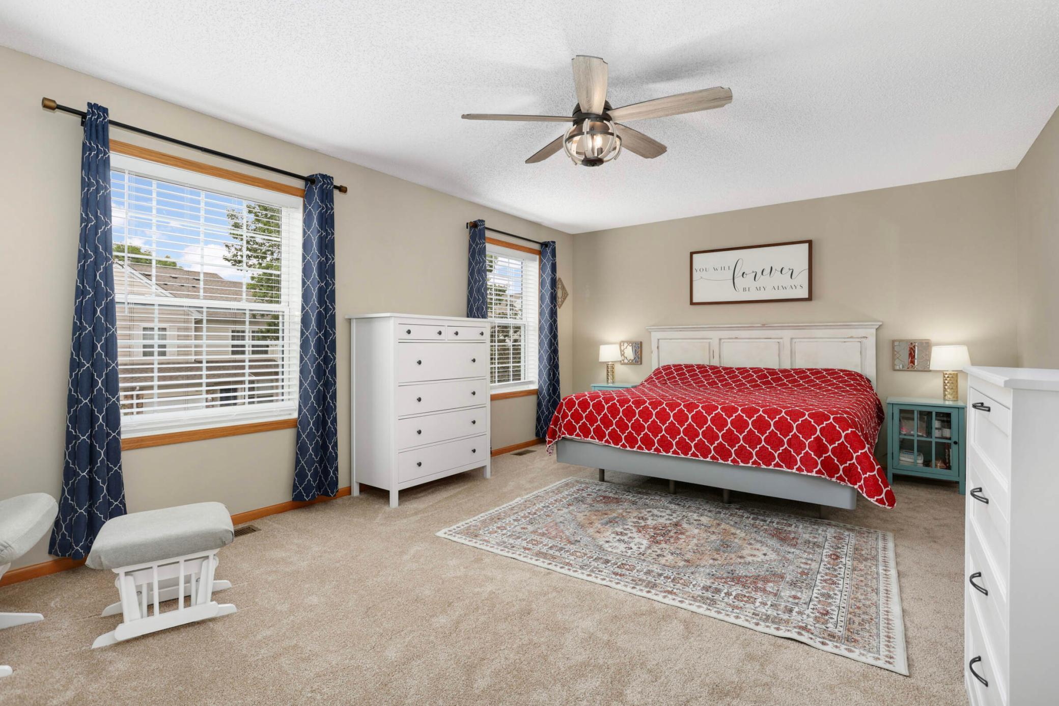 Bedroom with a red patterned bedspread, blue curtains, white dressers, and a ceiling fan.
