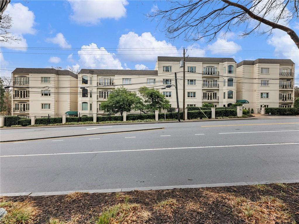 Cream-colored residential building with balconies behind a landscaped fence and roadside.