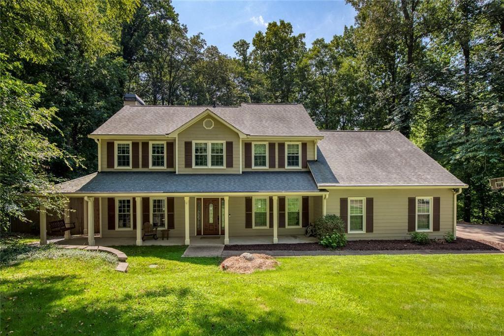 Two-story beige home with a covered porch, dark shutters, and a wooded lawn.