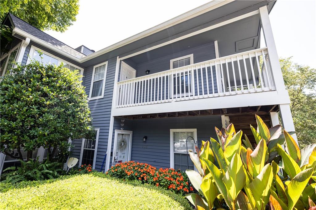 Blue two-story house with a white-railed balcony and lush flowering landscaping.