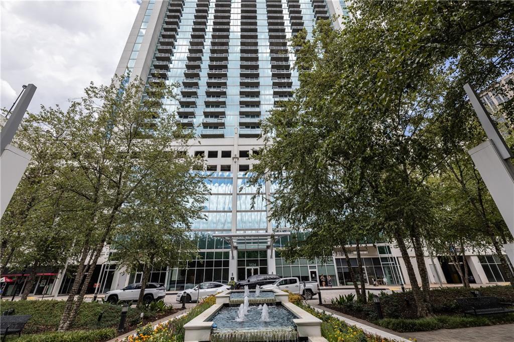 Modern high-rise entrance framed by trees, with a fountain in the landscaped forecourt.