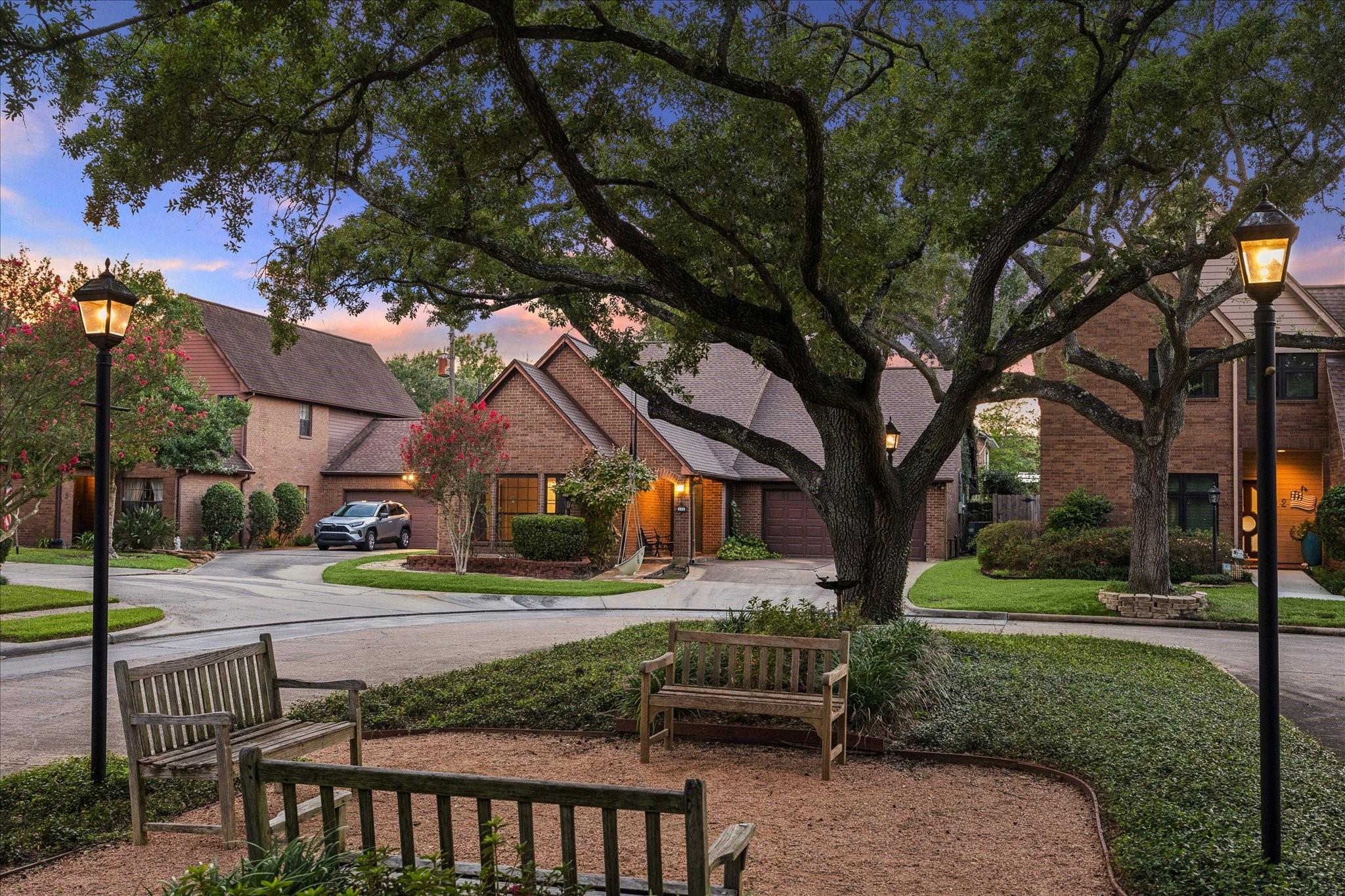 Brick homes beneath a sprawling oak beside wooden benches and glowing lampposts at dusk.