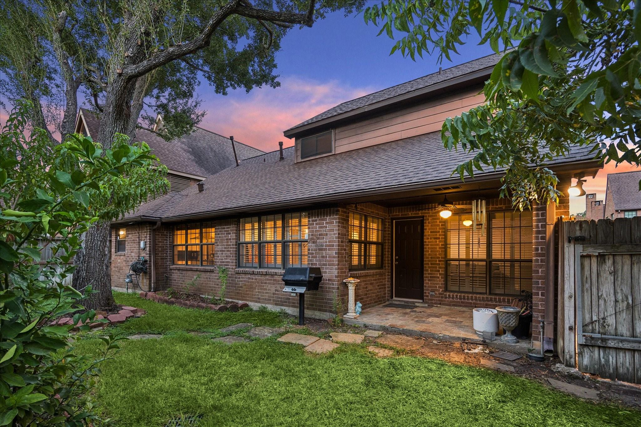 Brick home’s rear patio and lawn at dusk, framed by mature trees.