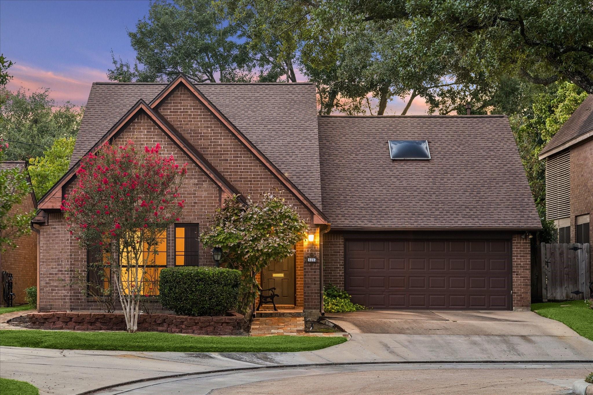 Brick house with steep gables, attached garage, and flowering trees at dusk.
