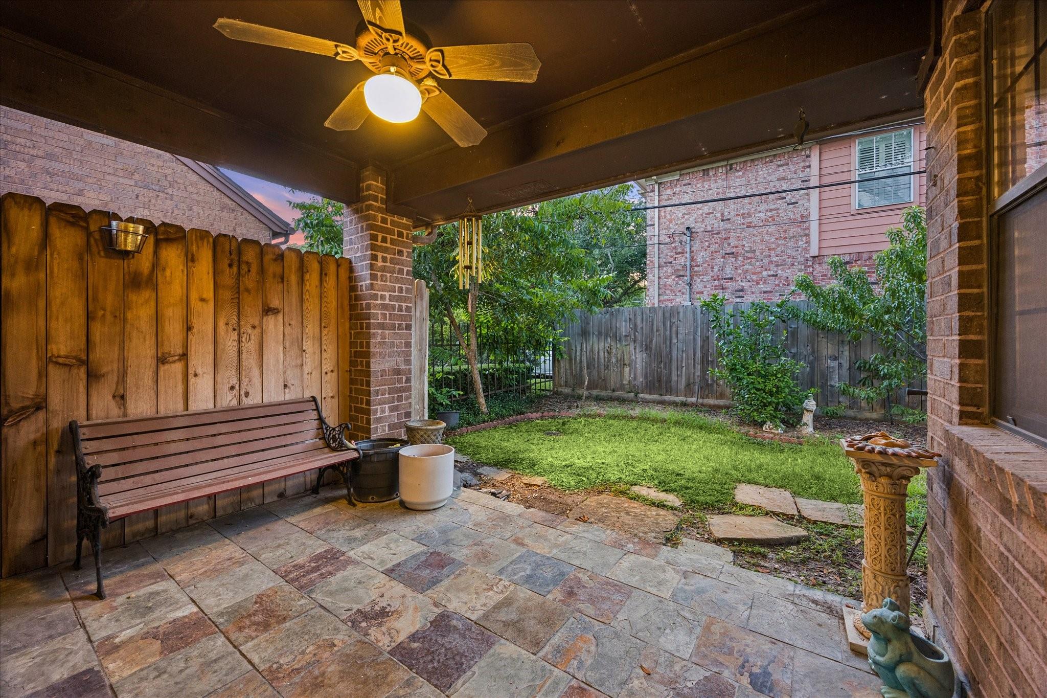 Covered stone patio with a wooden bench, ceiling fan, and small fenced lawn.