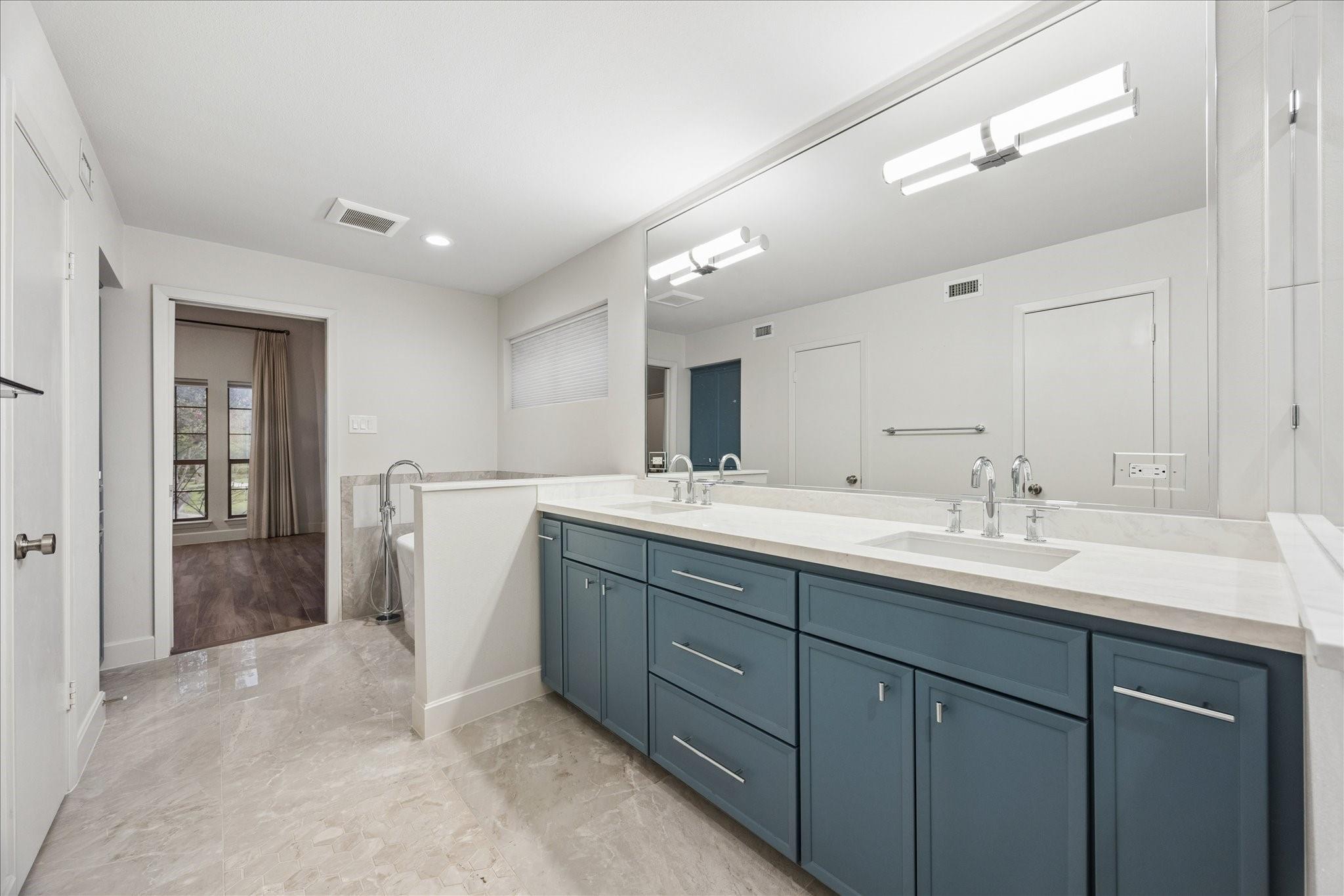 Bathroom with a long blue-gray double vanity, white countertop, large mirror, and stone tile floor.
