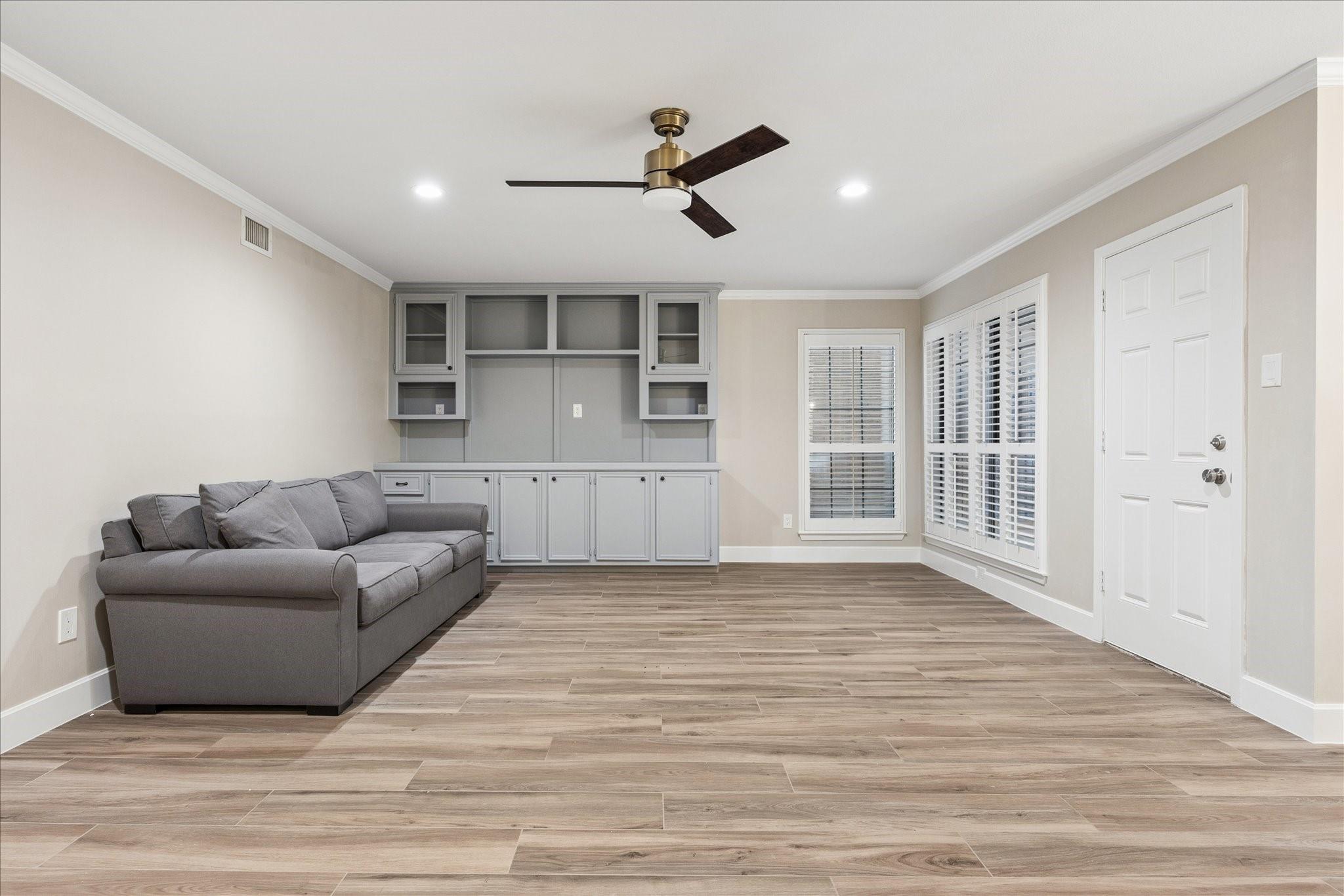 Living room with gray sofa, built-in cabinetry, wood-look tile flooring, and plantation shutters.