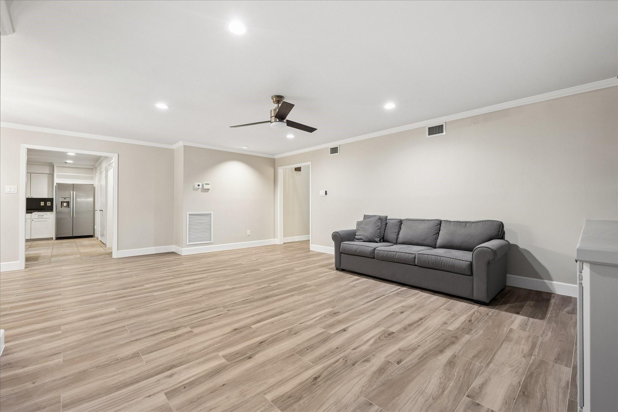Living room with a gray sofa, wood-look floors, ceiling fan, and open kitchen doorway.