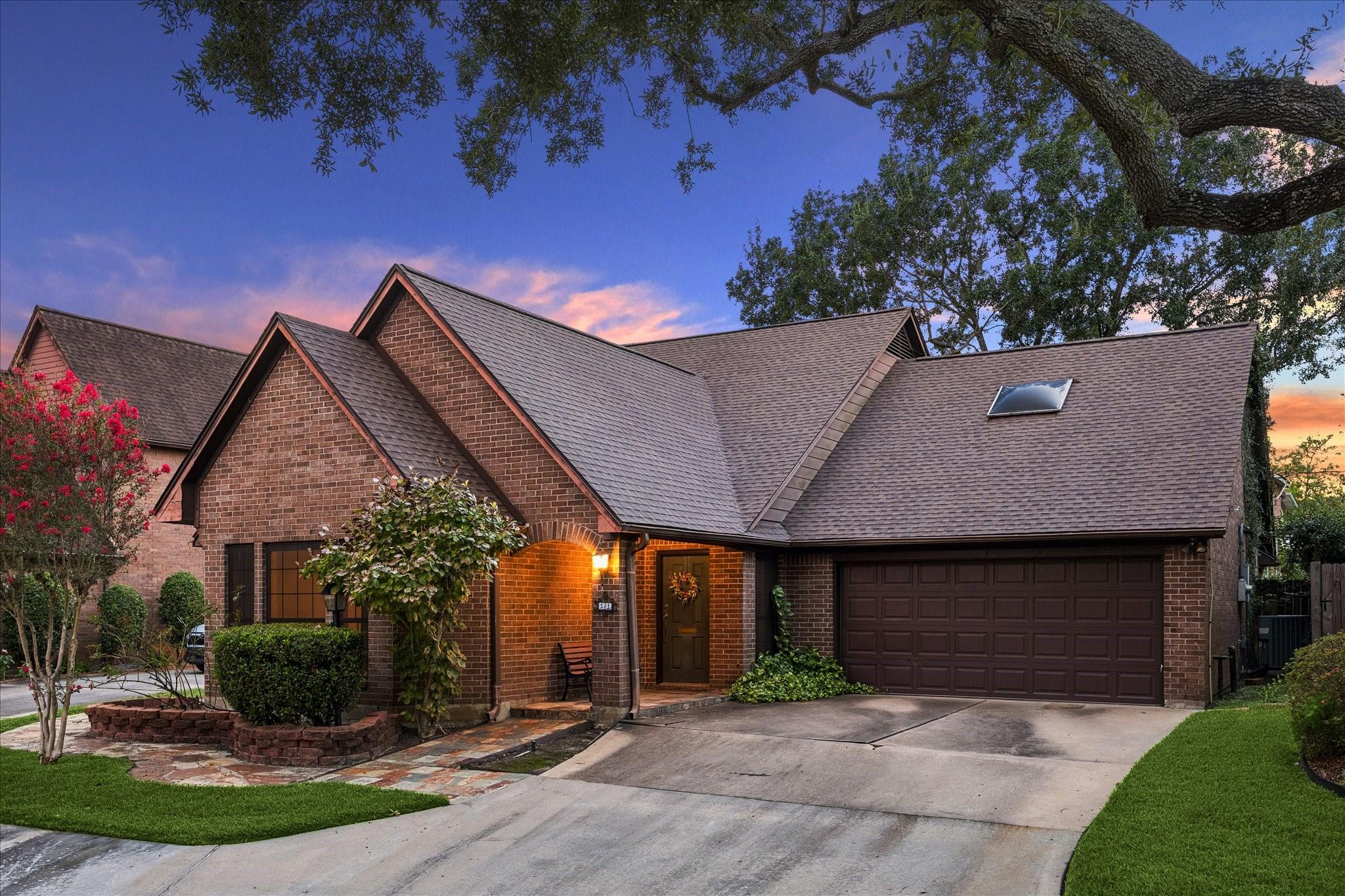 Brick house with a two-car garage and landscaped garden beneath a large oak tree at dusk.