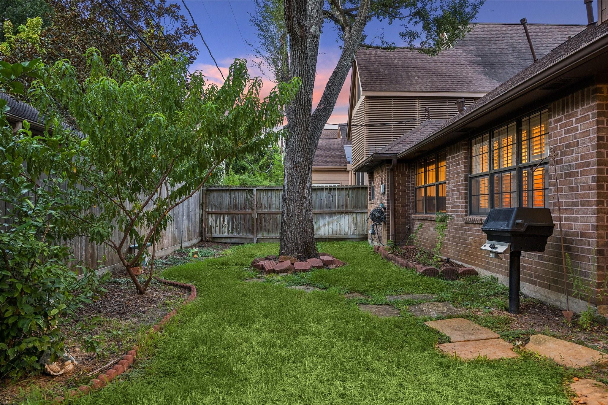 Fenced backyard with a broad lawn, mature tree, brick house, and barbecue grill at dusk.
