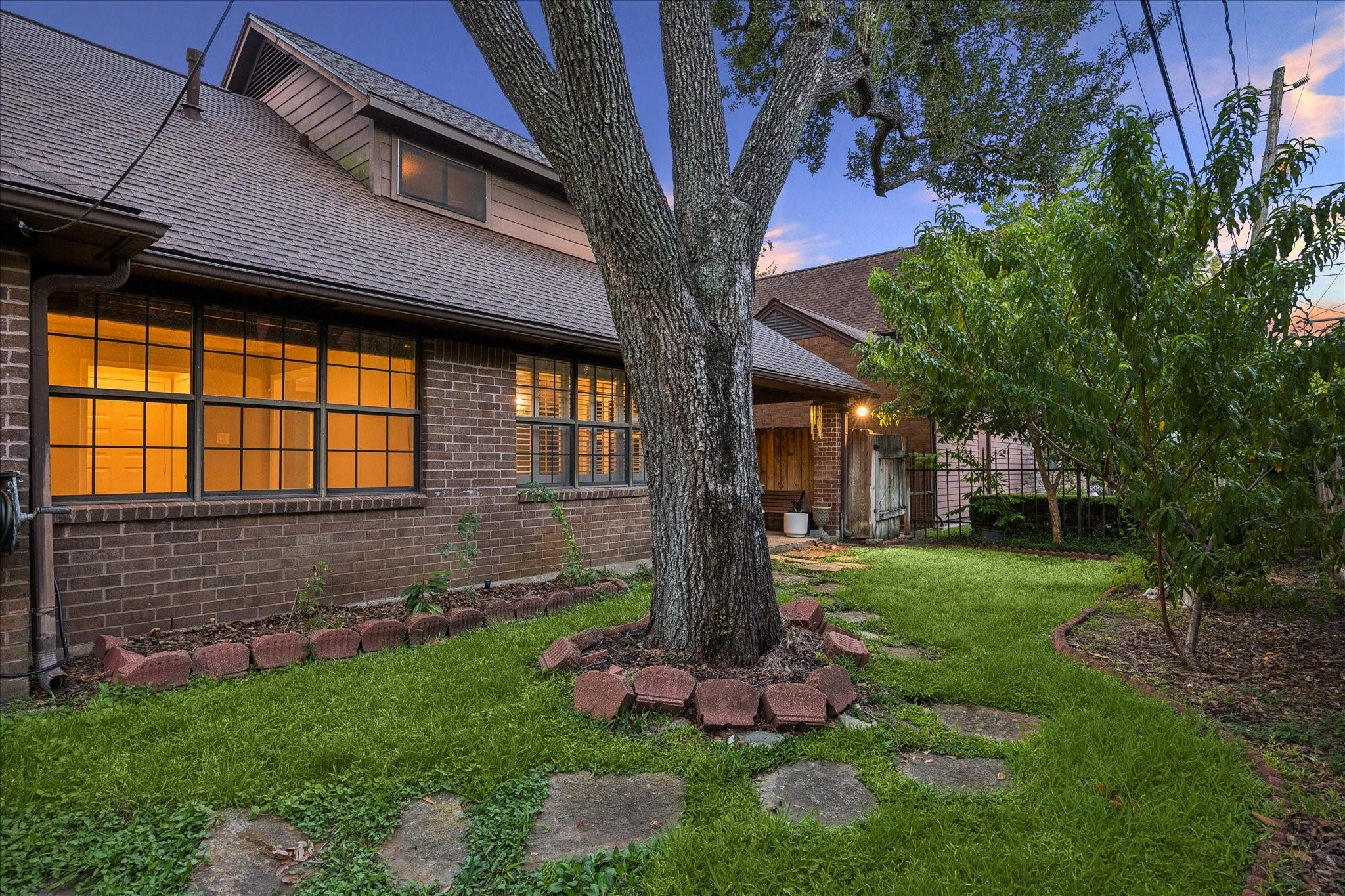 Brick house and backyard at dusk, with a large shade tree, glowing windows, and a stone path.