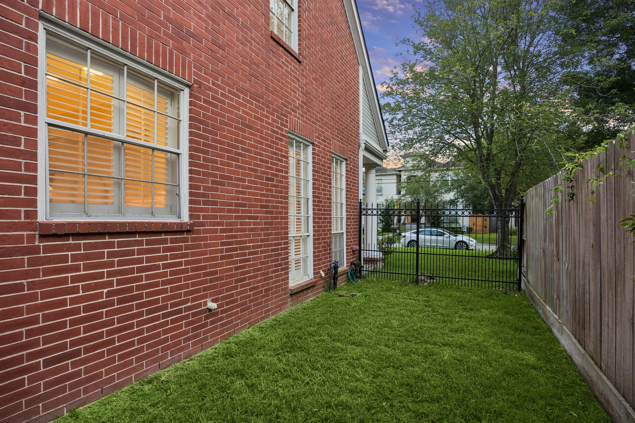 Side yard beside a red brick house, with a grassy lawn, black metal gate, and wooden fence.