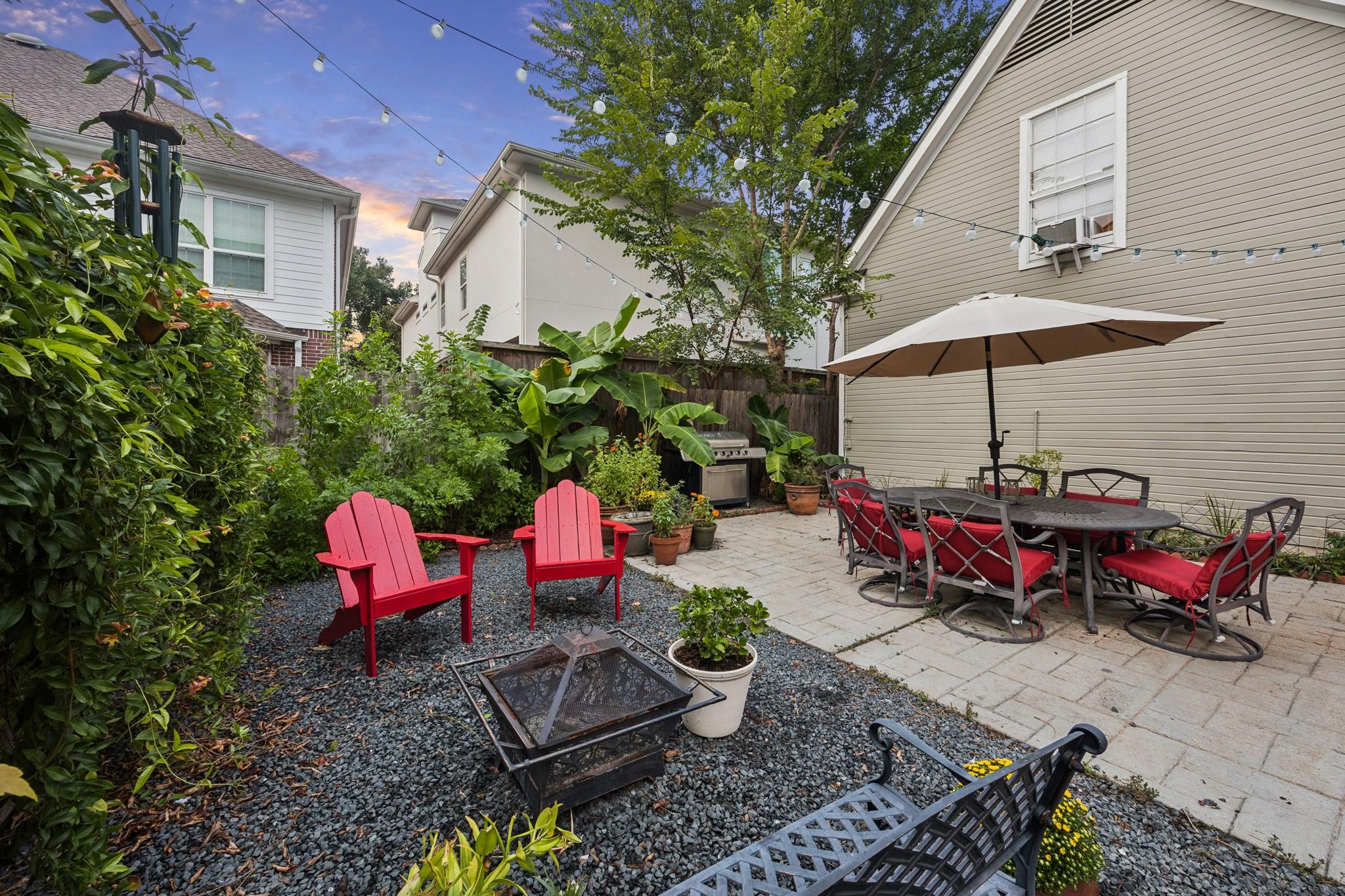 Backyard patio with umbrella-covered dining set, red Adirondack chairs, fire pit, and lush planting.