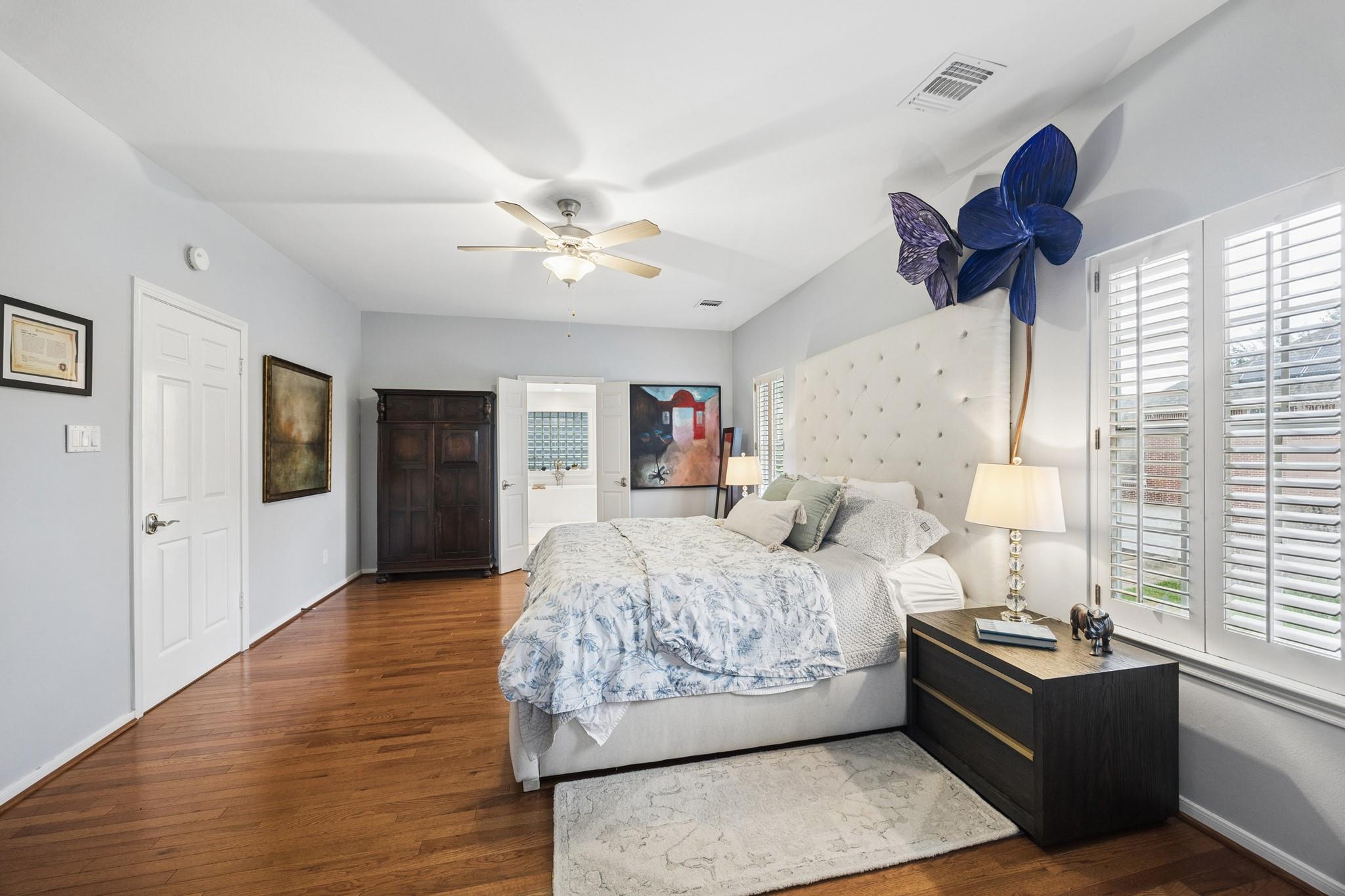 Bedroom with a tall tufted headboard, blue flower wall sculpture, plantation shutters, and hardwood floors.