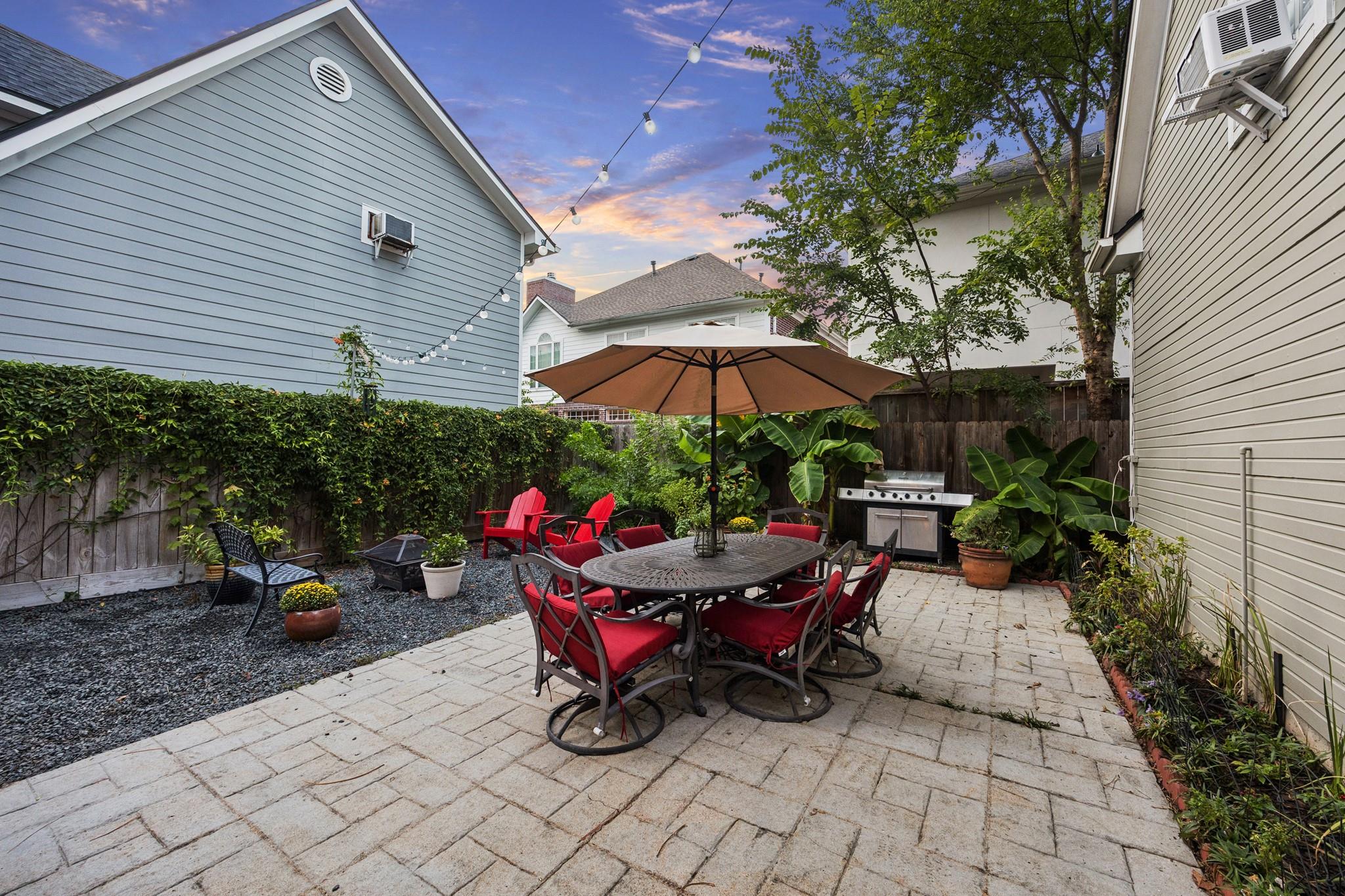 Backyard patio with umbrella-shaded dining table, red chairs, barbecue grill, and string lights.