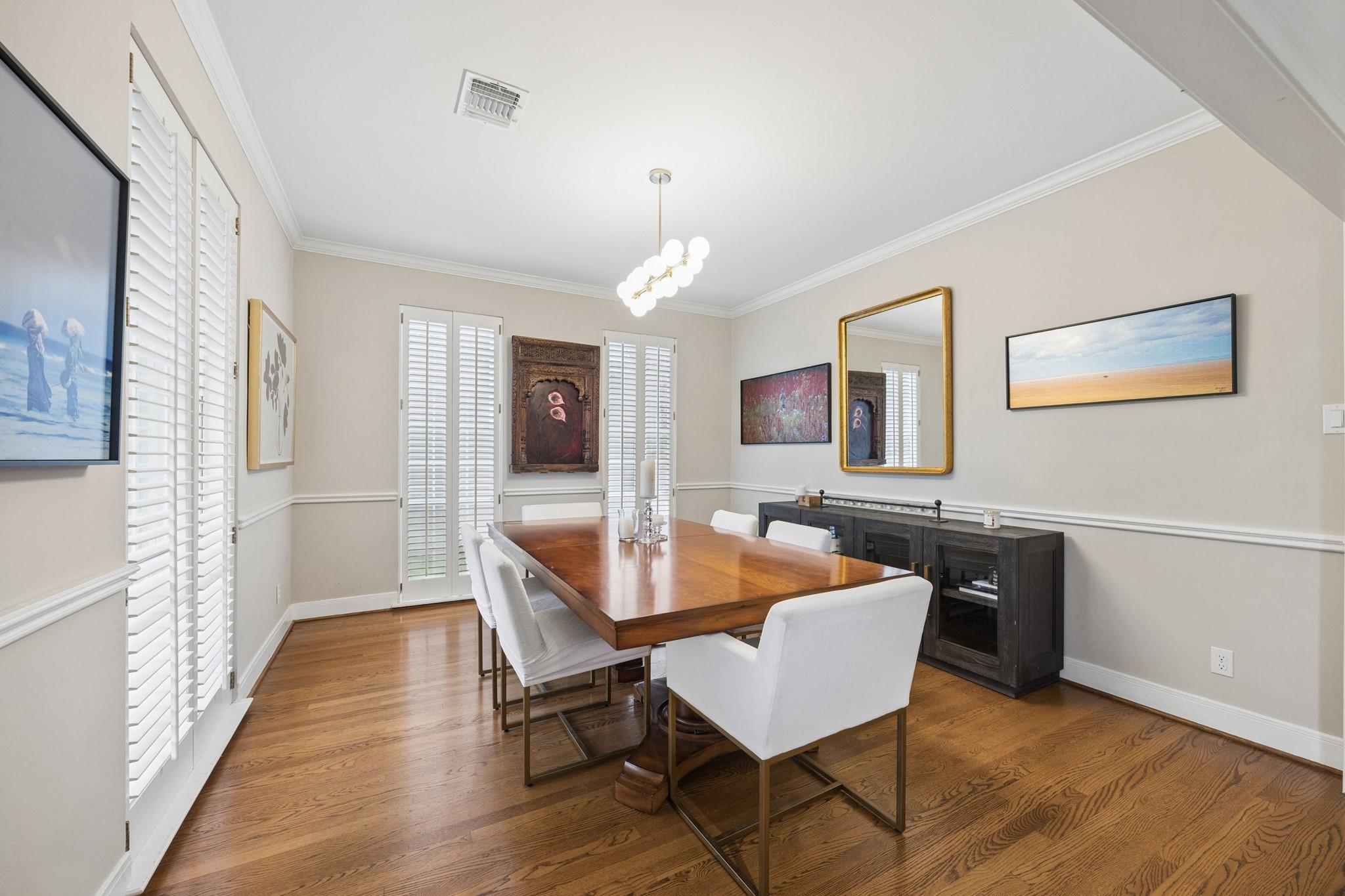 Dining room with a long wooden table, white chairs, hardwood floors, and plantation shutters.